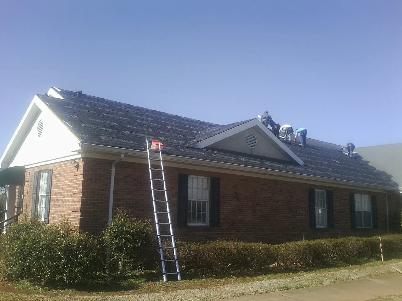 Crew tearing shingles off a brick ranch roof with a ladder set — Metal Roof Repair work by our crew in Dexter